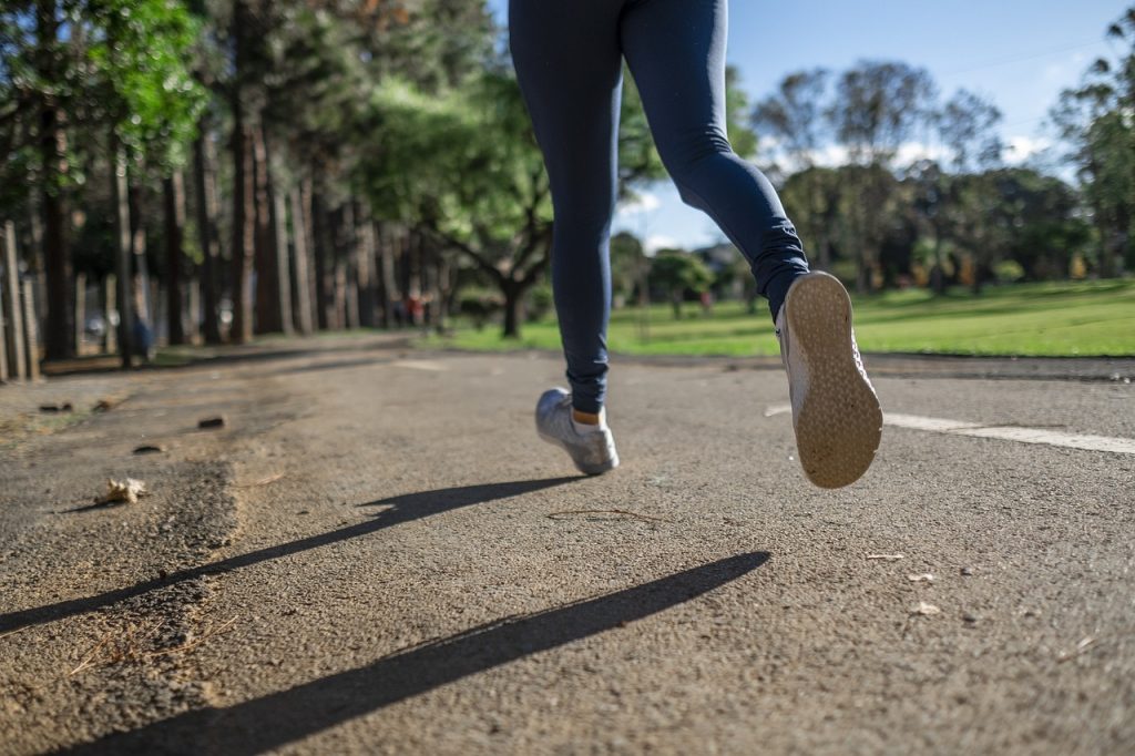 A person jogging in a park, captured in mid-stride with a focus on their blue leggings and the soles of their running shoes, amidst a blurred background of trees and a walking path